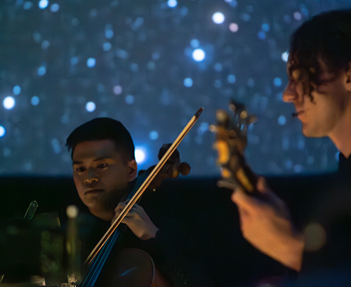 A photo of a cellist and a guitarist playing in the darkness, with a wall of blue stars behind them.