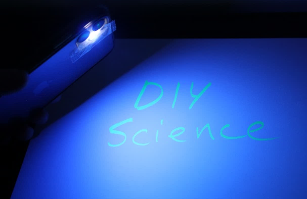 A dark blue page with a phone shining a light on the words "DIY Science" which appear to be glowing in UV light.