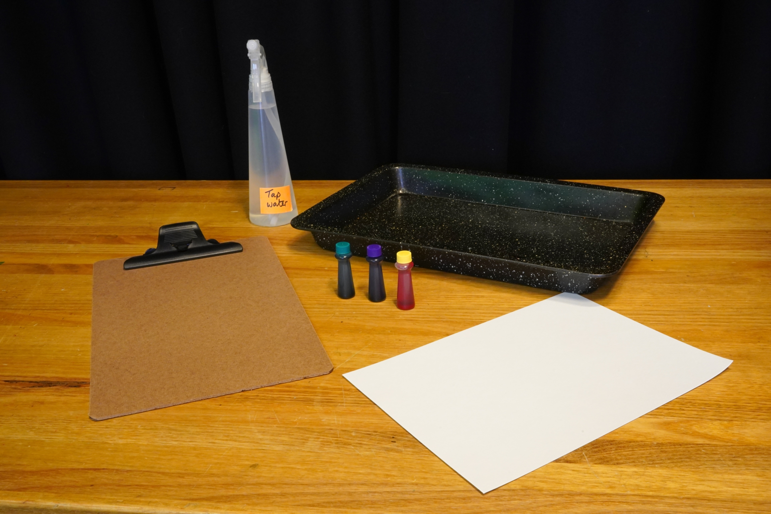 A picture of the materials, a clipboard, a white piece of paper, a baking tray, a spray bottle and 3 different food colouring droppers.